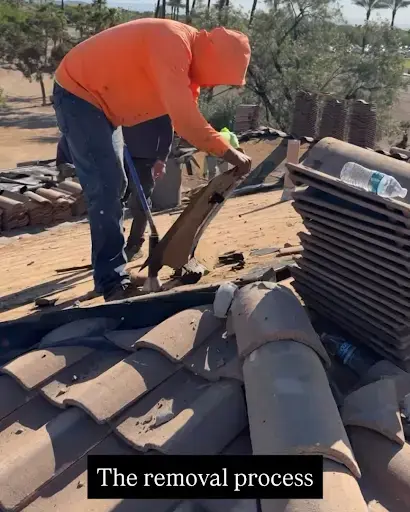 damaged-underlayment | Triumph Roofing Worker in an orange hoodie removes clay roof tiles on a pitched roof during a removal process, with tiles scattered nearby.