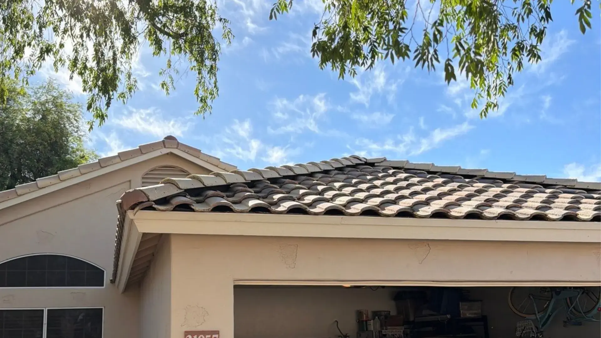roofing-repair-in-glendale-az | Triumph Roofing Beige house with a curved clay tile roof, an open garage showing shelves and a bicycle, under a bright blue sky with overhanging branches of trees.